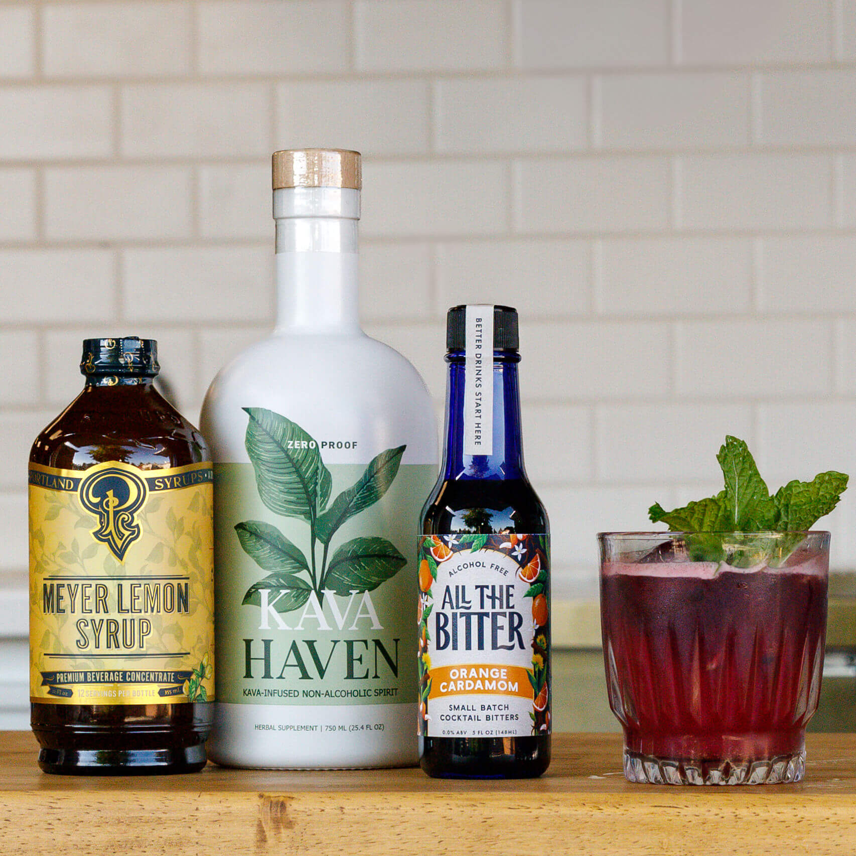 Bottles of Kava Haven, Meyer Lemon Syrup, All The Bitter, and a mocktail with a glass on a wooden surface.