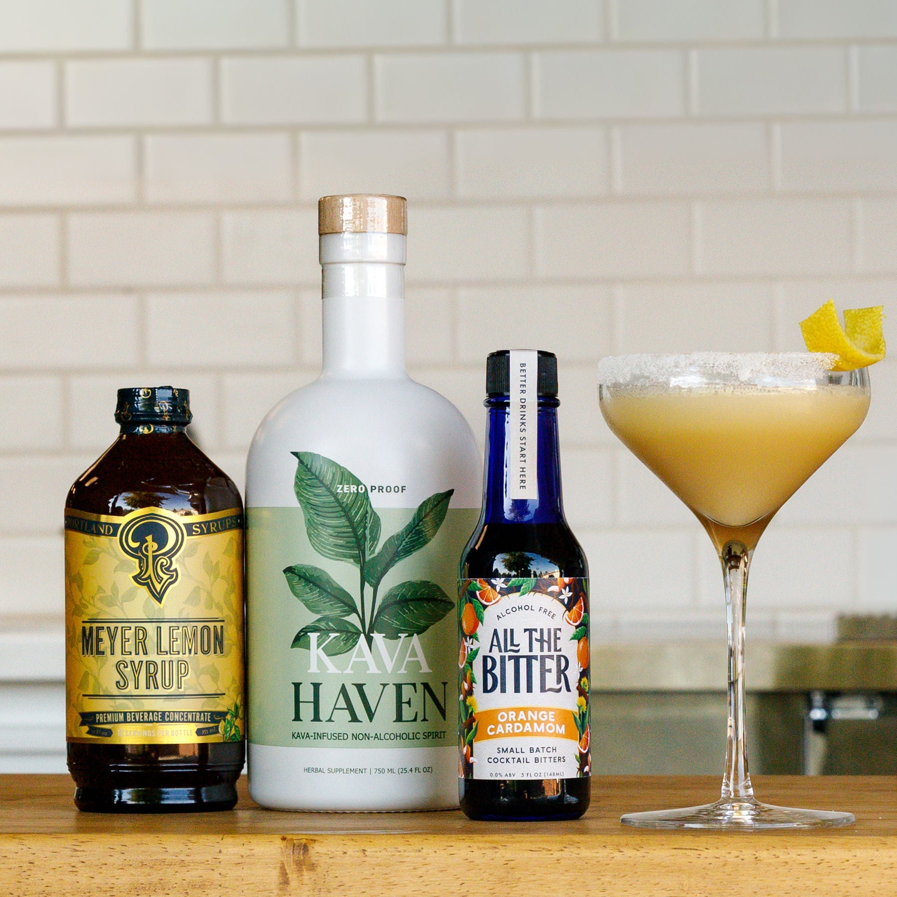 Three bottles of Kava Haven, Meyer Lemon Syrup, and All The Bitter on a wooden surface with a cocktail in the background.