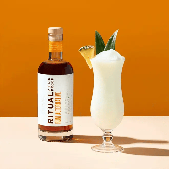 Bottle of Ritual Zero Proof Rum Alternative with a cocktail garnished with pineapple on an orange background
